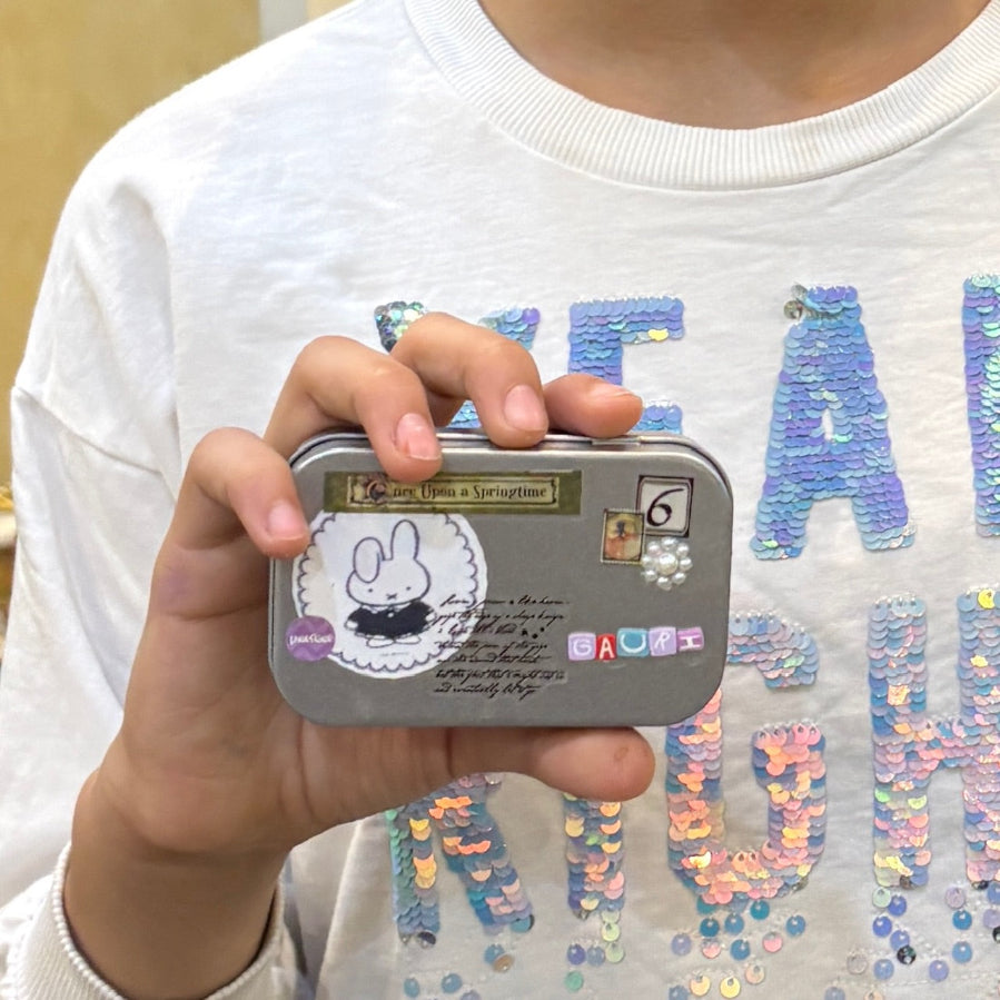 Hand holding a small tin with a rabbit design, against a white shirt with colorful text.