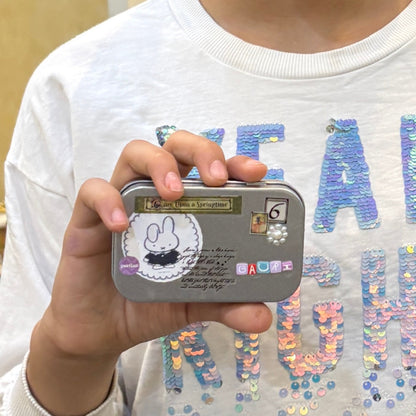 Hand holding a small tin with a rabbit design, against a white shirt with colorful text.