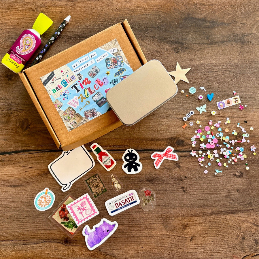 Contents of the kit on a table. It consists of a box with a tin, a variety of stickers and assorted embellishments to be applied with a wax pencil and glue.