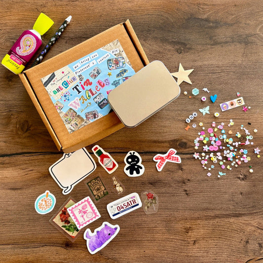 Contents of the kit on a table. It consists of a box with a tin, a variety of stickers and assorted embellishments to be applied with a wax pencil and glue.