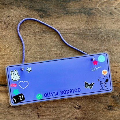 Blue rectangular sign with decorative stickers and 'Olivia Rodrigo' text on a wooden surface